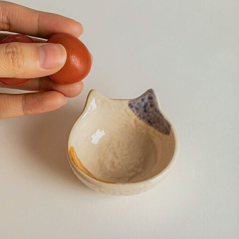 Close-up of a cream ceramic cat-shaped dipping dish shown beside a tomato.
